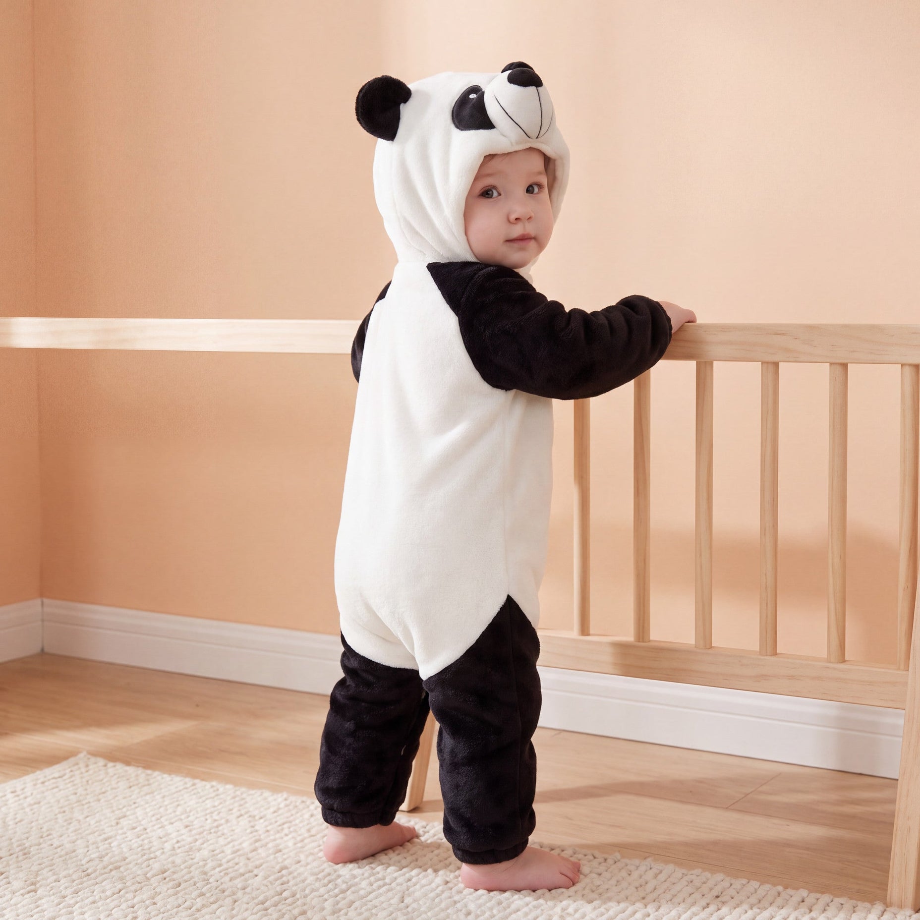 Panda one-piece on child in a supported standing pose in a peach nursery, attached panda hood and open ankle cuffs.